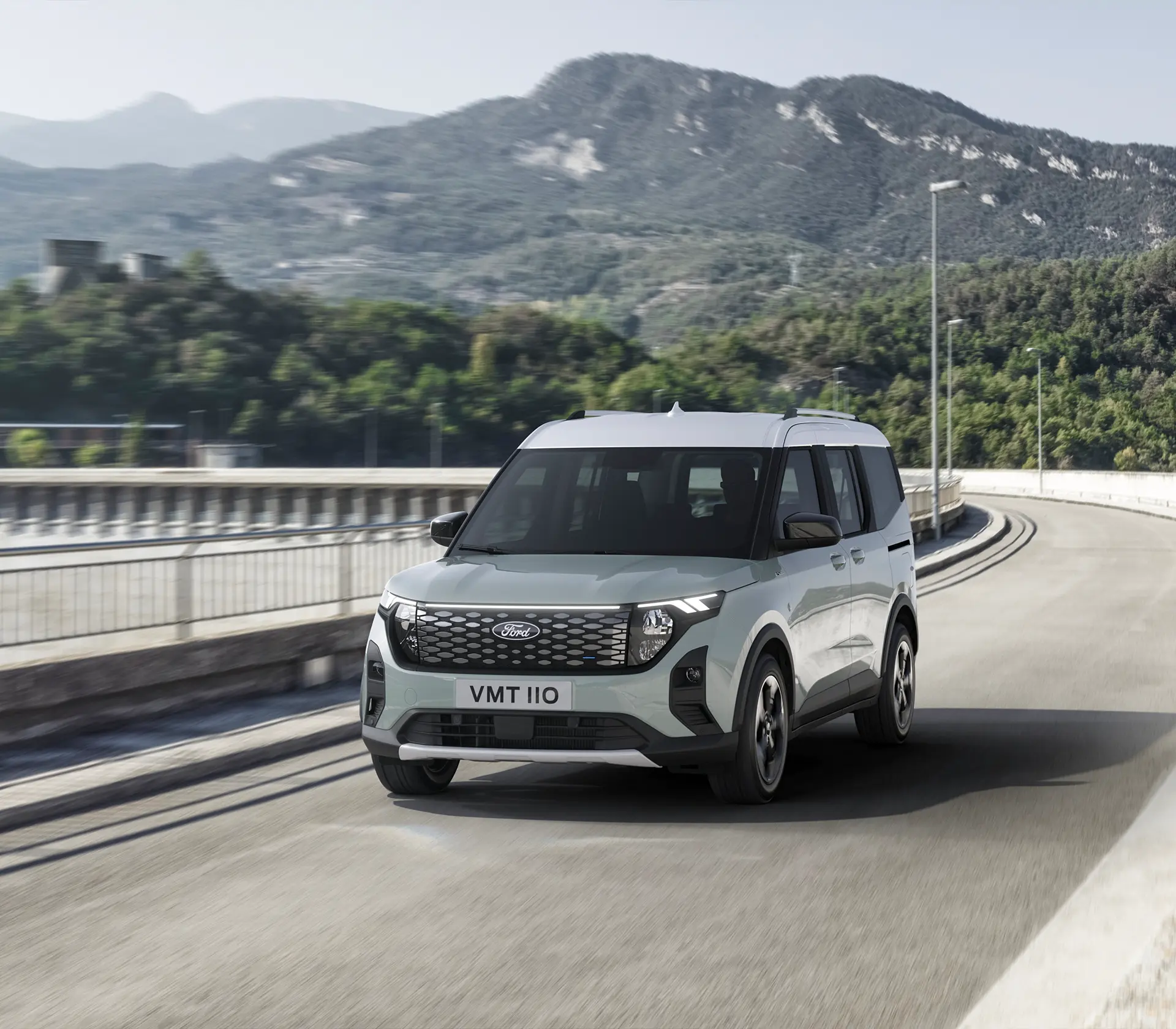 Grauer E-Tourneo Courier fährt auf der Straße mit Gebirgen im Hintergrund