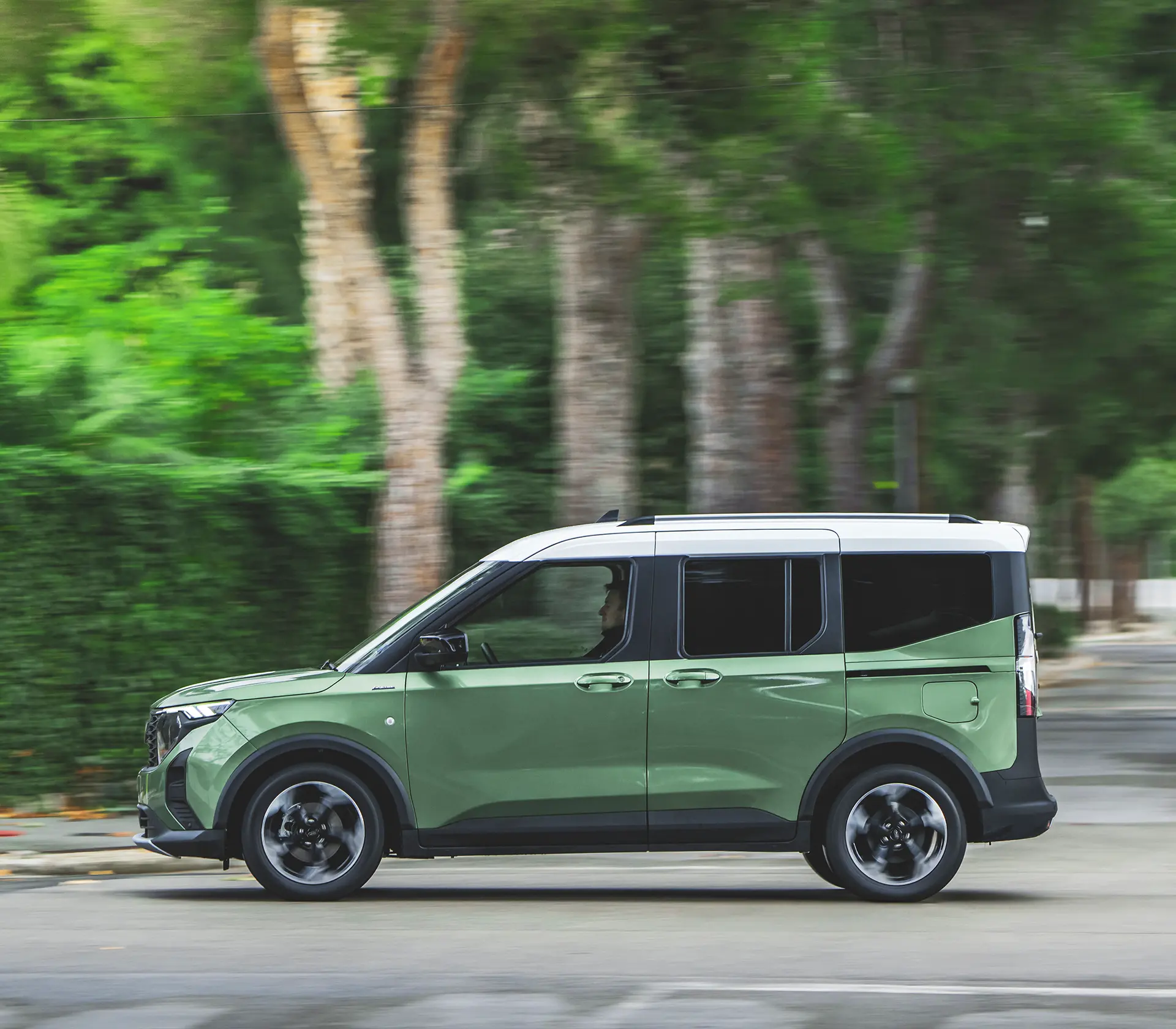 eine Seitliche Aufnahme des Grünen Tourneo Courier welcher auf einer Straße fährt mit Bäumen im Hintergrund