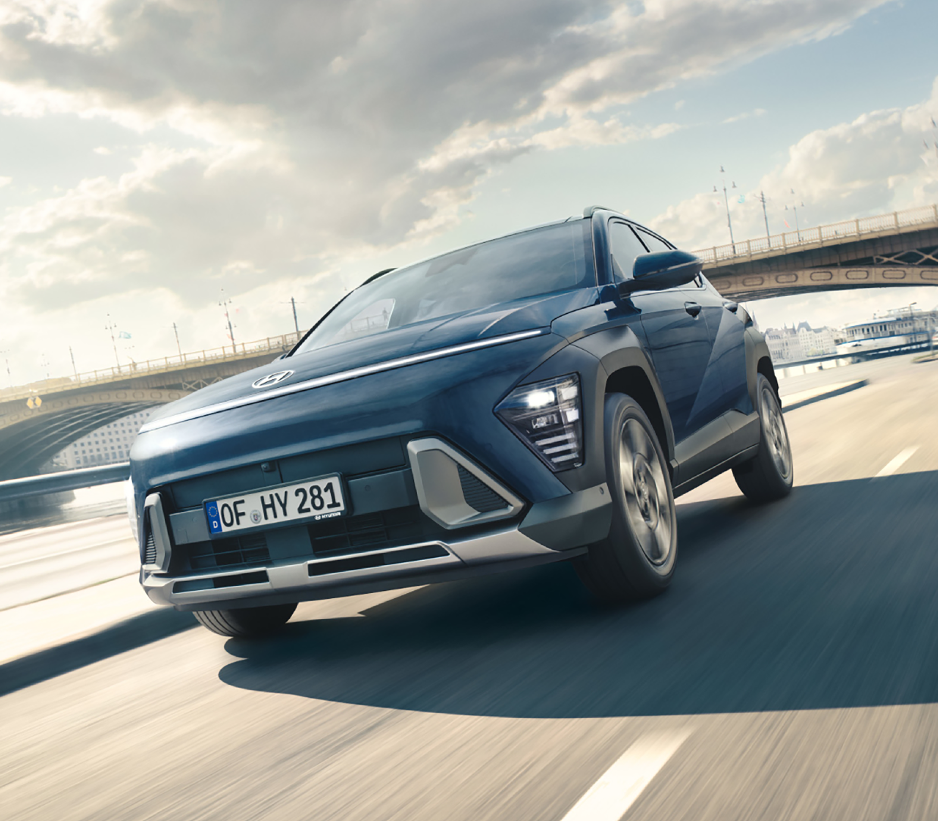Hyundai KONA fährt auf der Straße mit einer Brücke und Himmel mit Wolken im Hintergrund