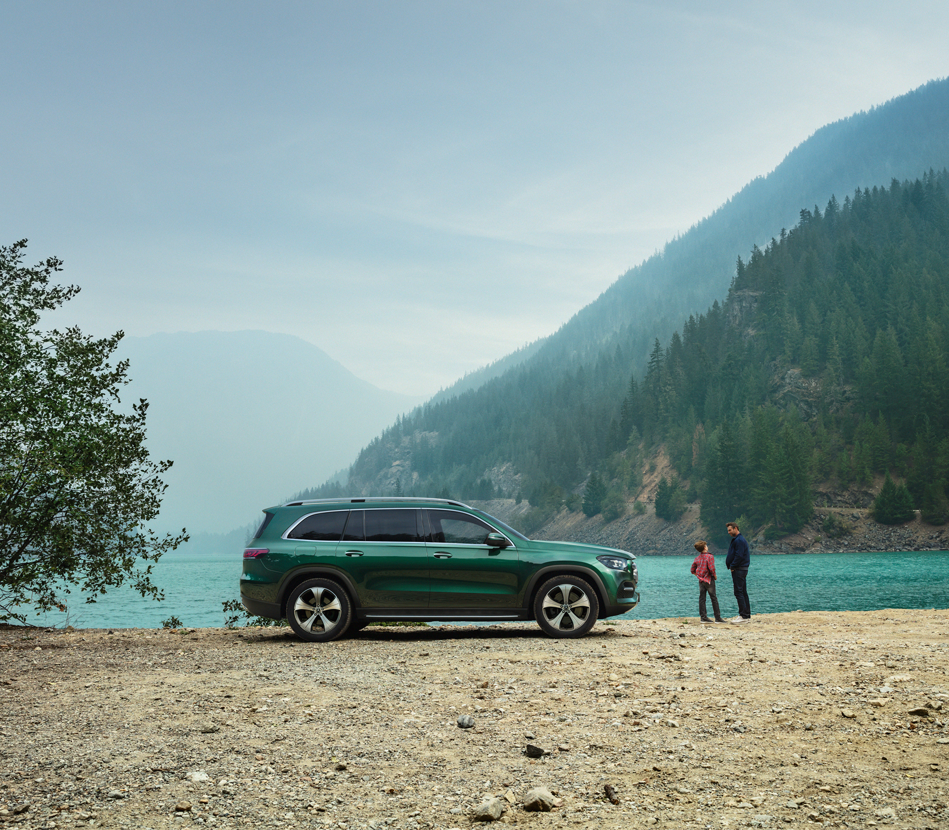 Mercedes-Benz GLS Seitenansicht und von der Seite steht geparkt vor einem See mit Bäumen und Bergen im Hintergrund, man sieht zwei Menschen die vor dem Auto stehen