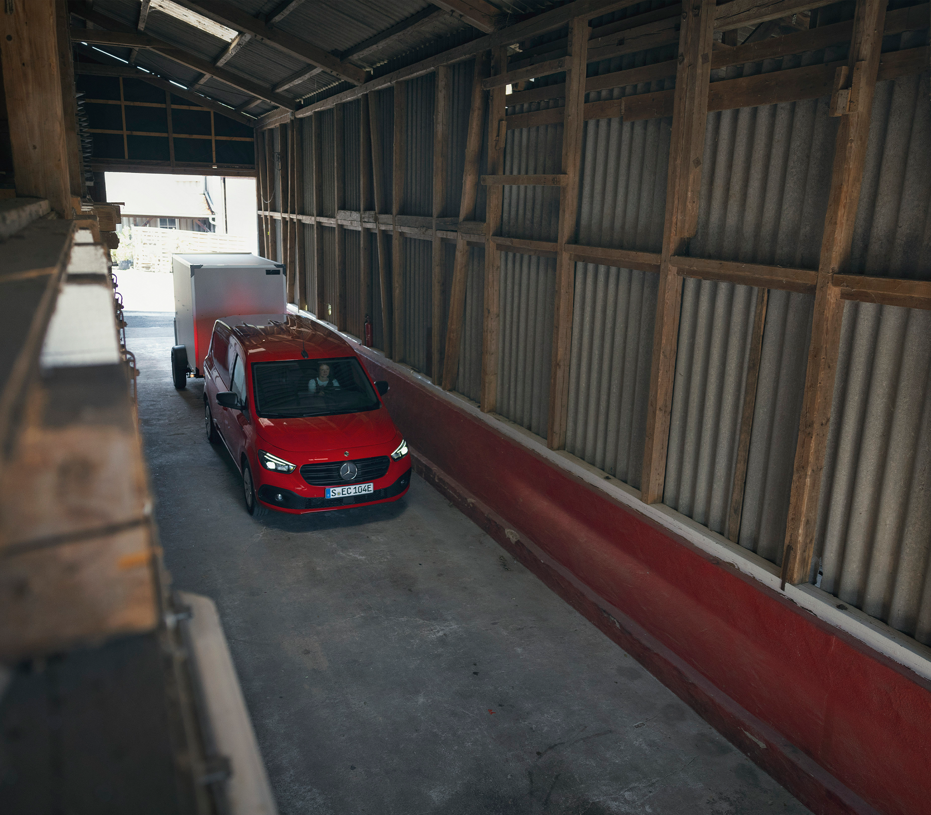 Roter Mercedes-Benz Citan fährt durch eine enge Halle mit Holz- und Blechverkleidung mit einem Anhänger