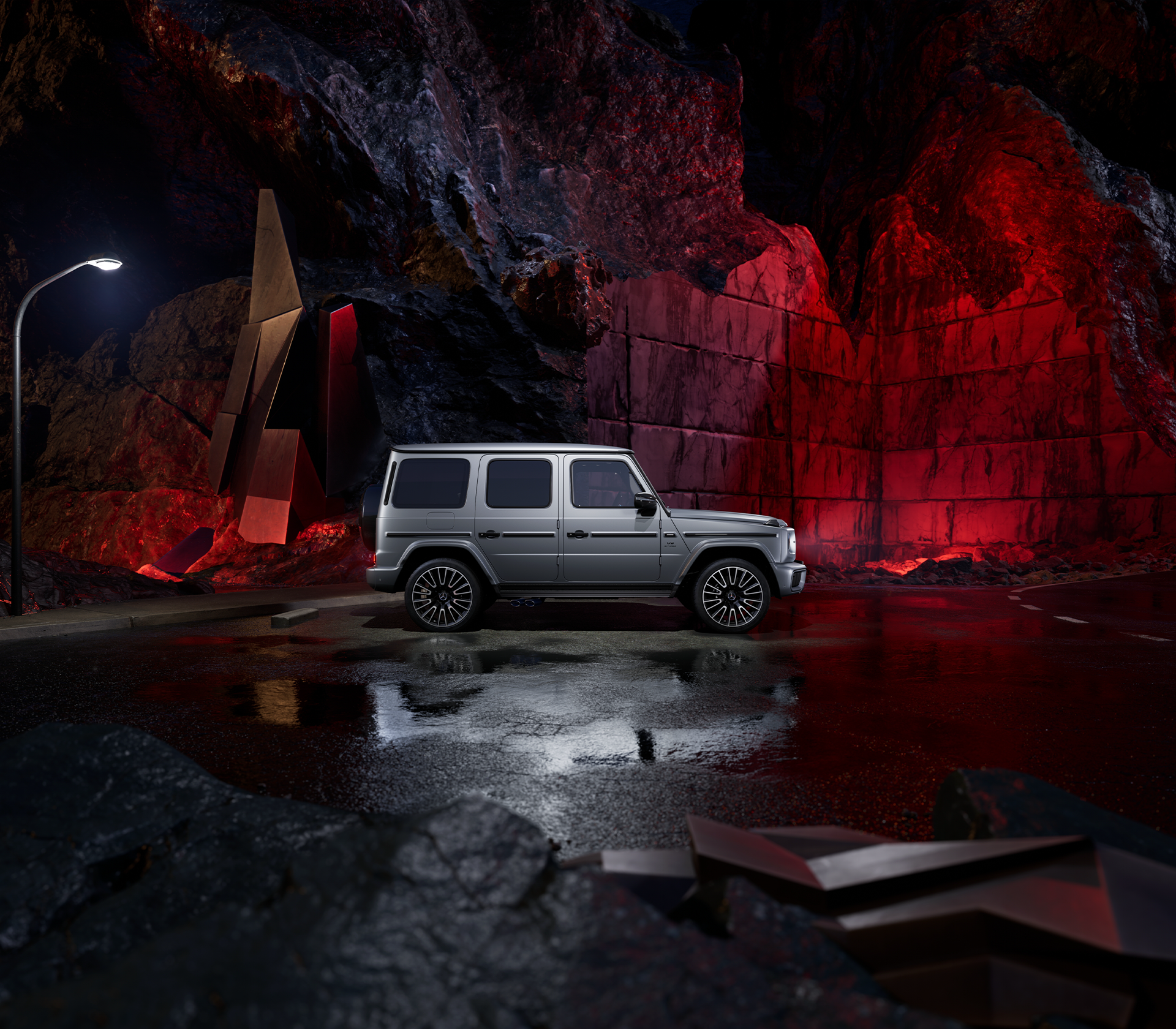 Seitliche Ansicht einer Mercedes-Benz G-Klasse in grau, inszeniert vor dramatischer Felskulisse mit roter Beleuchtung und nasser Fahrbahn.