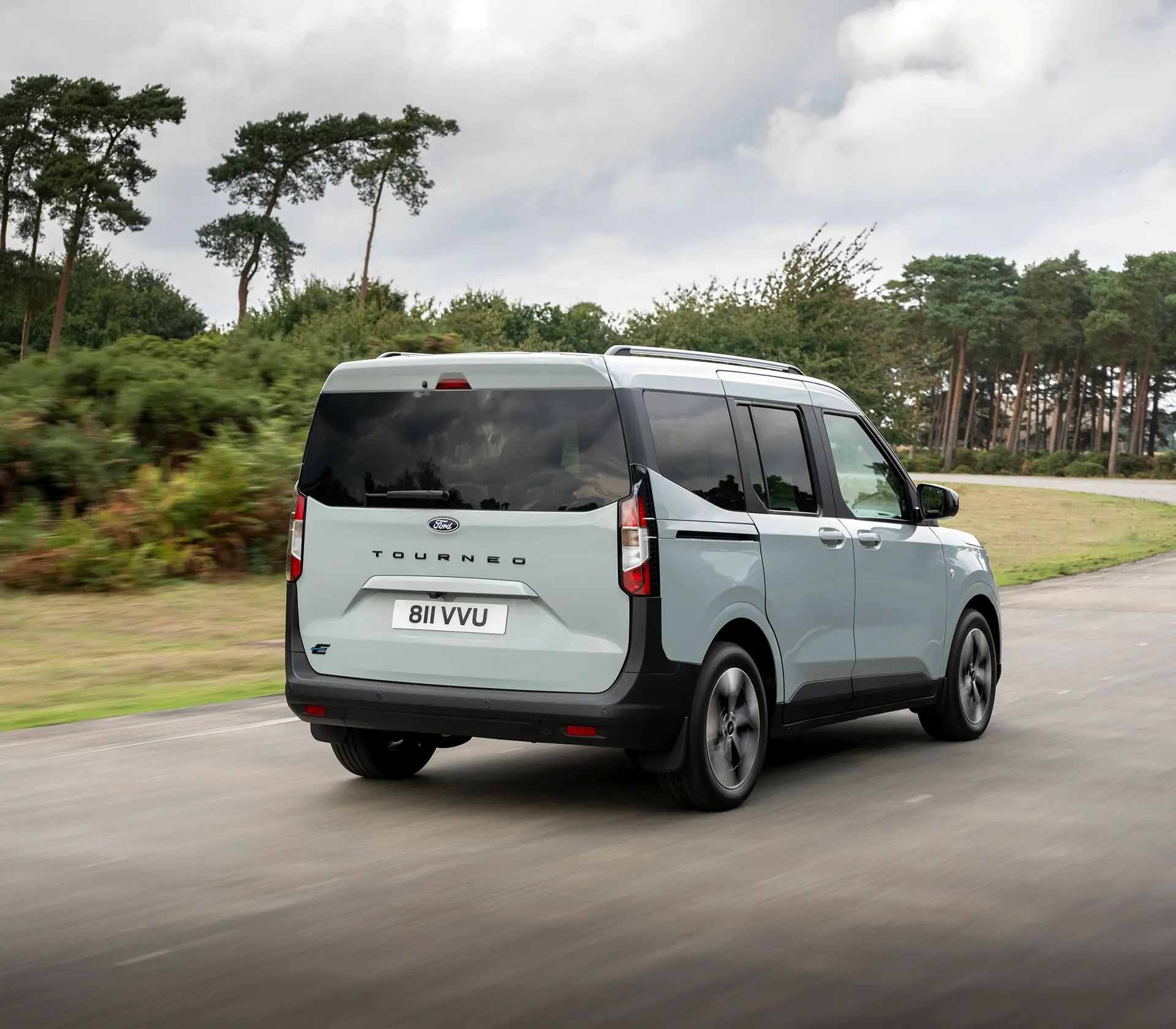 Grauer E-Tourneo Courier fährt auf der Straße mit Gebirgen im Hintergrund