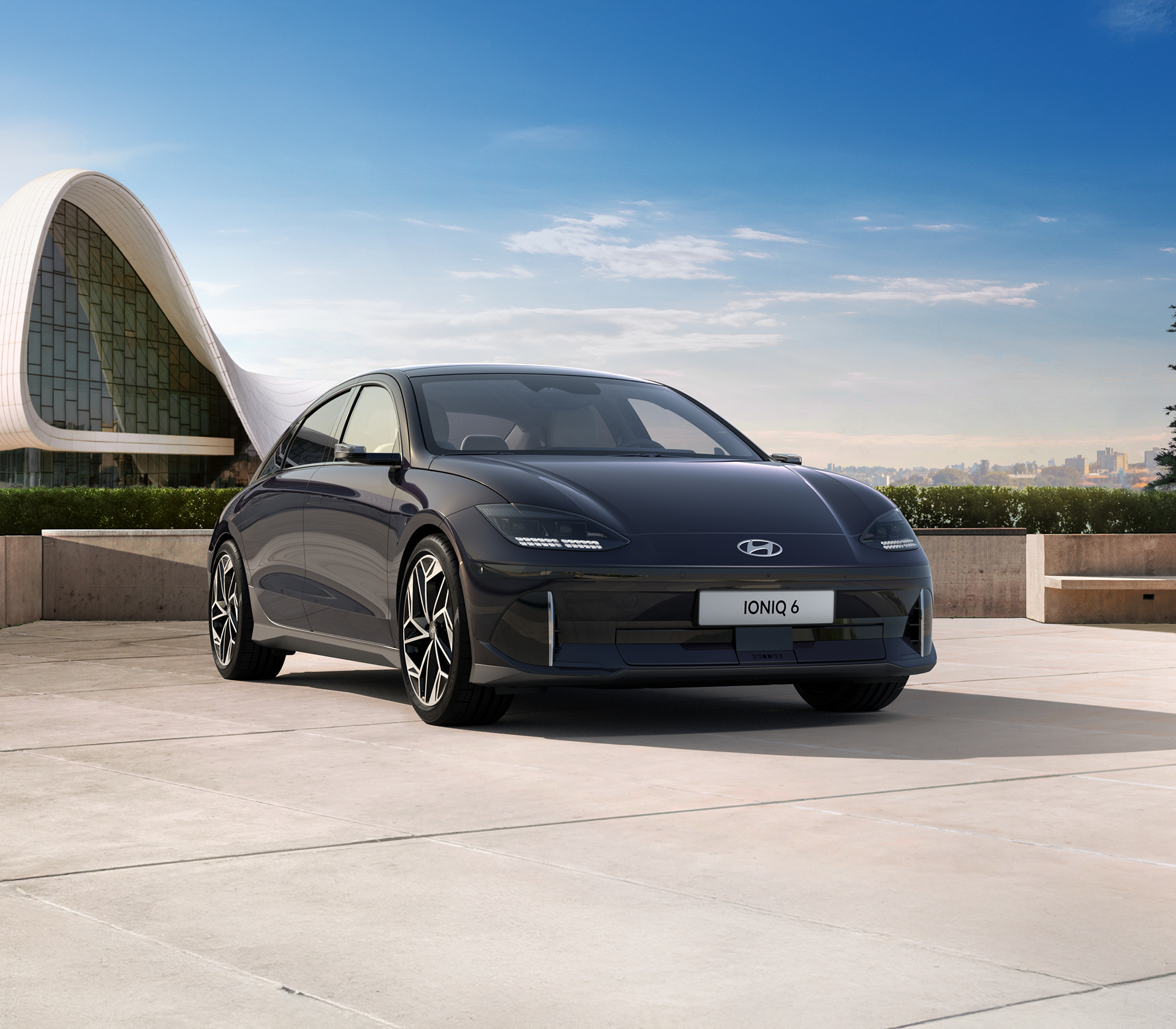 Hyundai IONIC 6 in schwarz, Hintergrund Himmel, Bäume und ein modernes Gebäude von vorne mit einem modernen Gebäude im Hintergrund und blauem Himmel