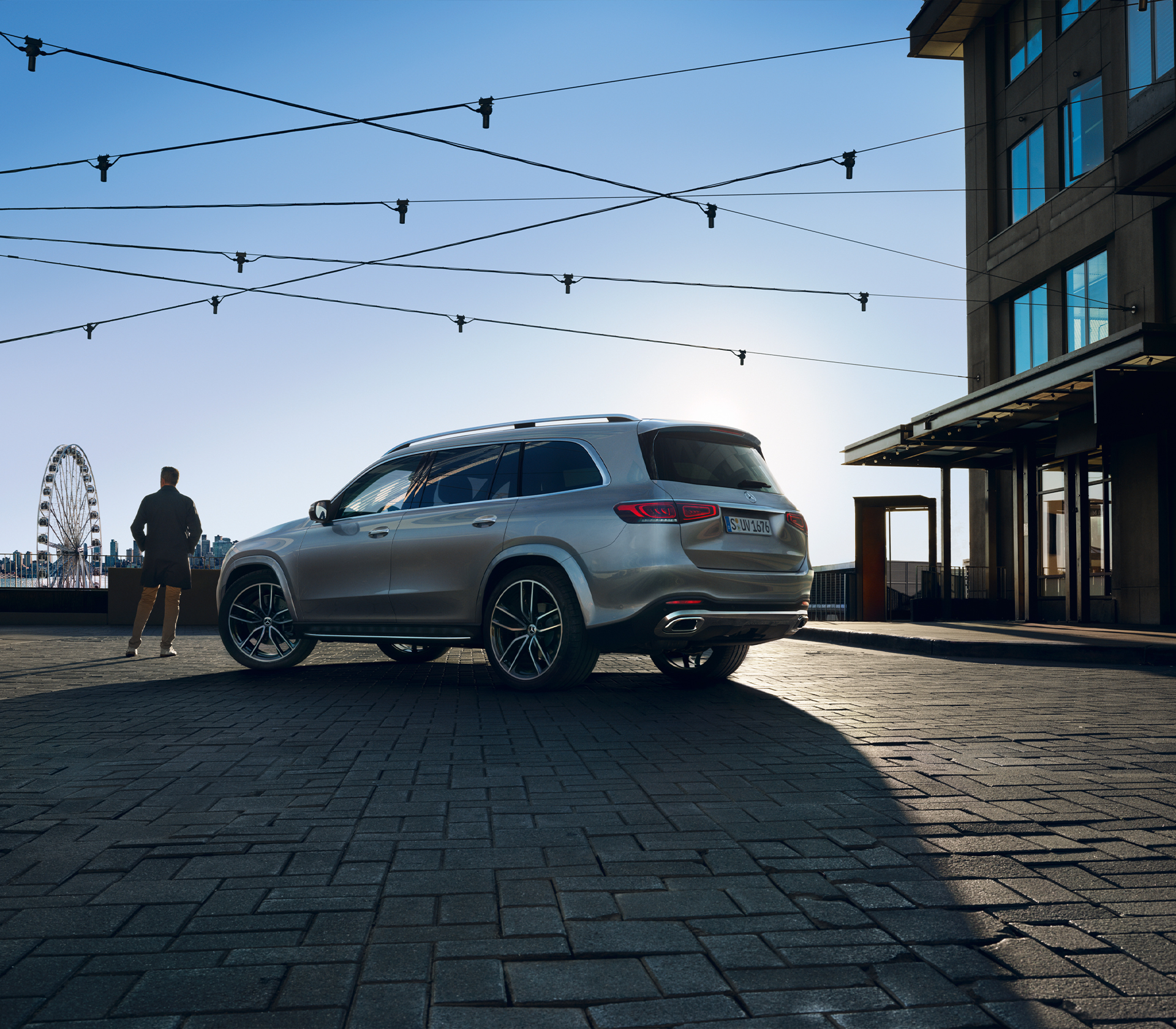 Mercedes-Benz GLS Seiten- und Heckansicht mit einer lichterkette drüber einem Haus auf der rechten seite einem Menschen und riesenrad im Hintergrund und der blaue Abendhimmel