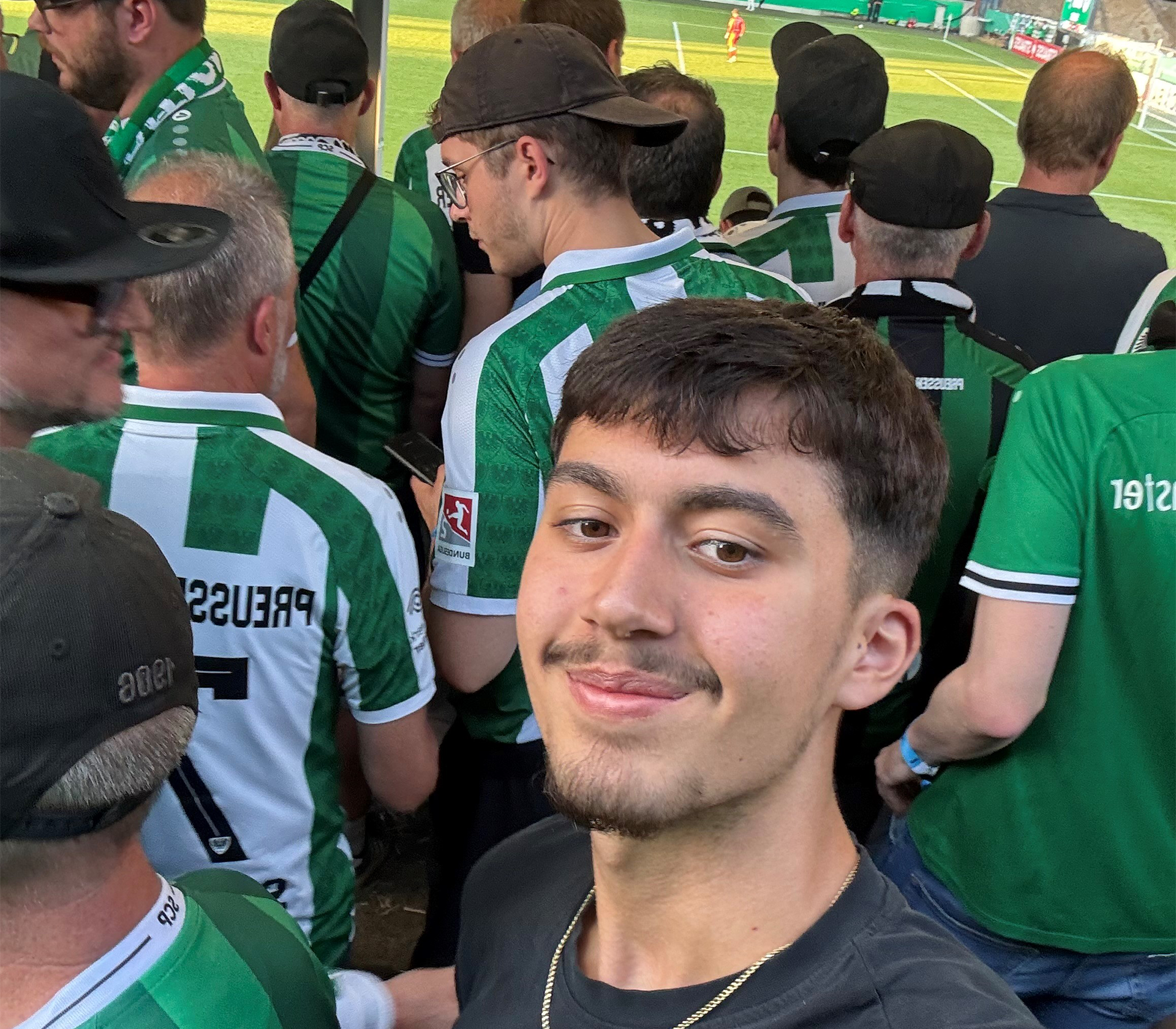 Ein Selfie von einem jungen Mann. Im Hintergrund sind Männer mit Trikots vom preußen Münster und einem Fußballfeld.