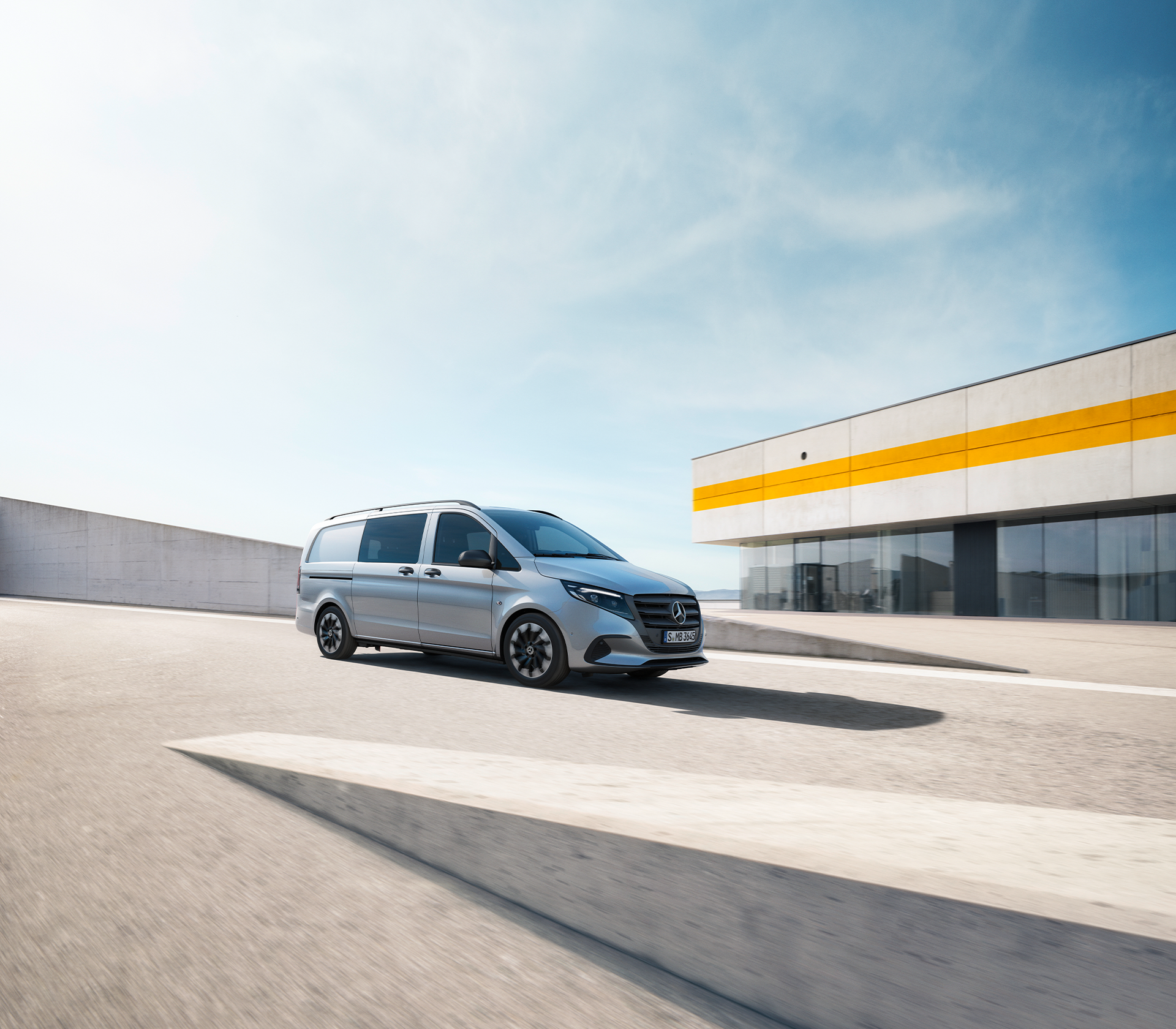 „Fahrender silberner Mercedes-Benz Vito vor moderner Industriearchitektur mit gelbem Streifendetail.“