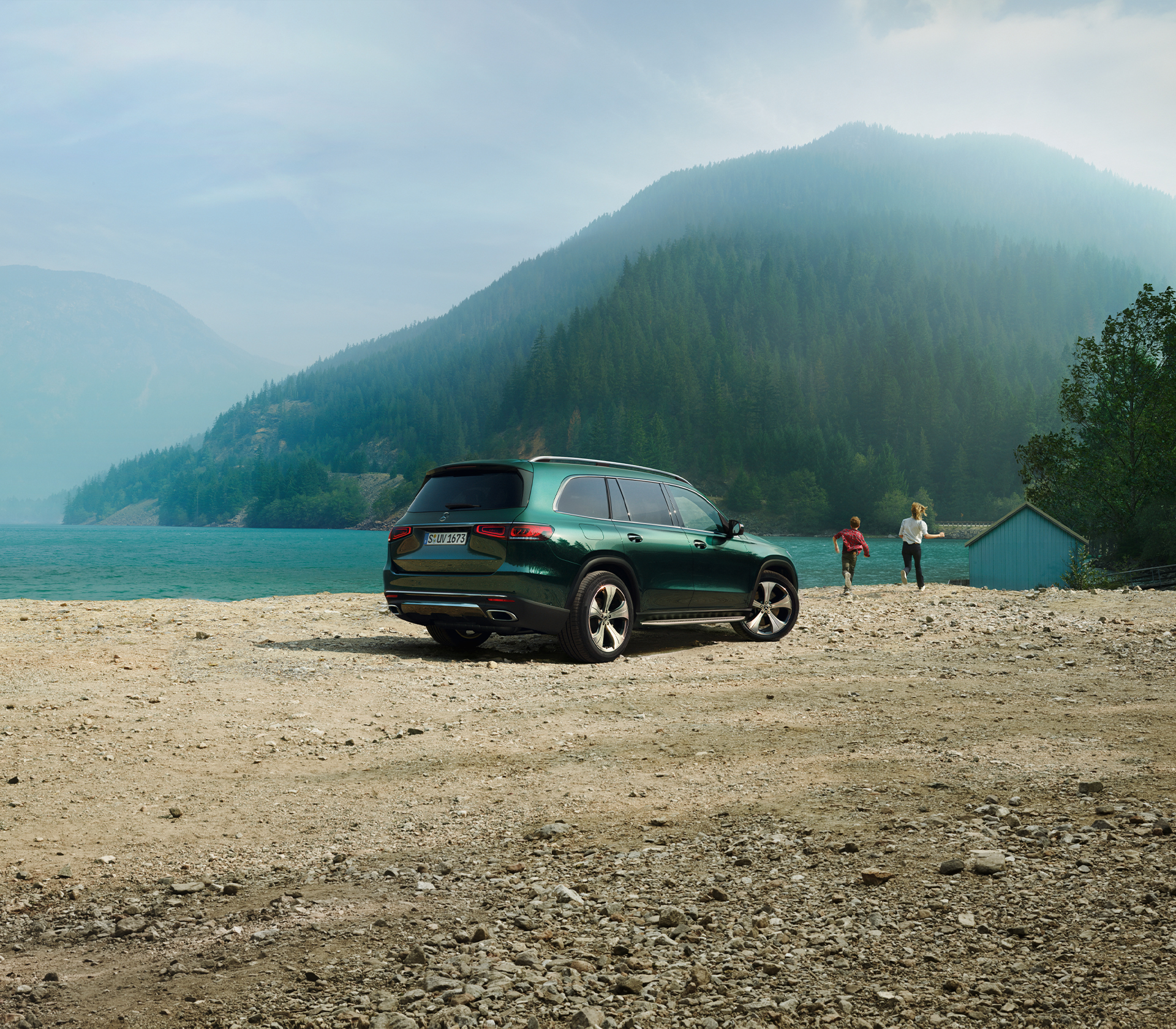 Mercedes-Benz GLS HEckansicht und von der Seite steht geparkt vor einem See mit Bäumen und Bergen im Hintergrund, man sieht zwei Menschen die zu einem Haus laufen
