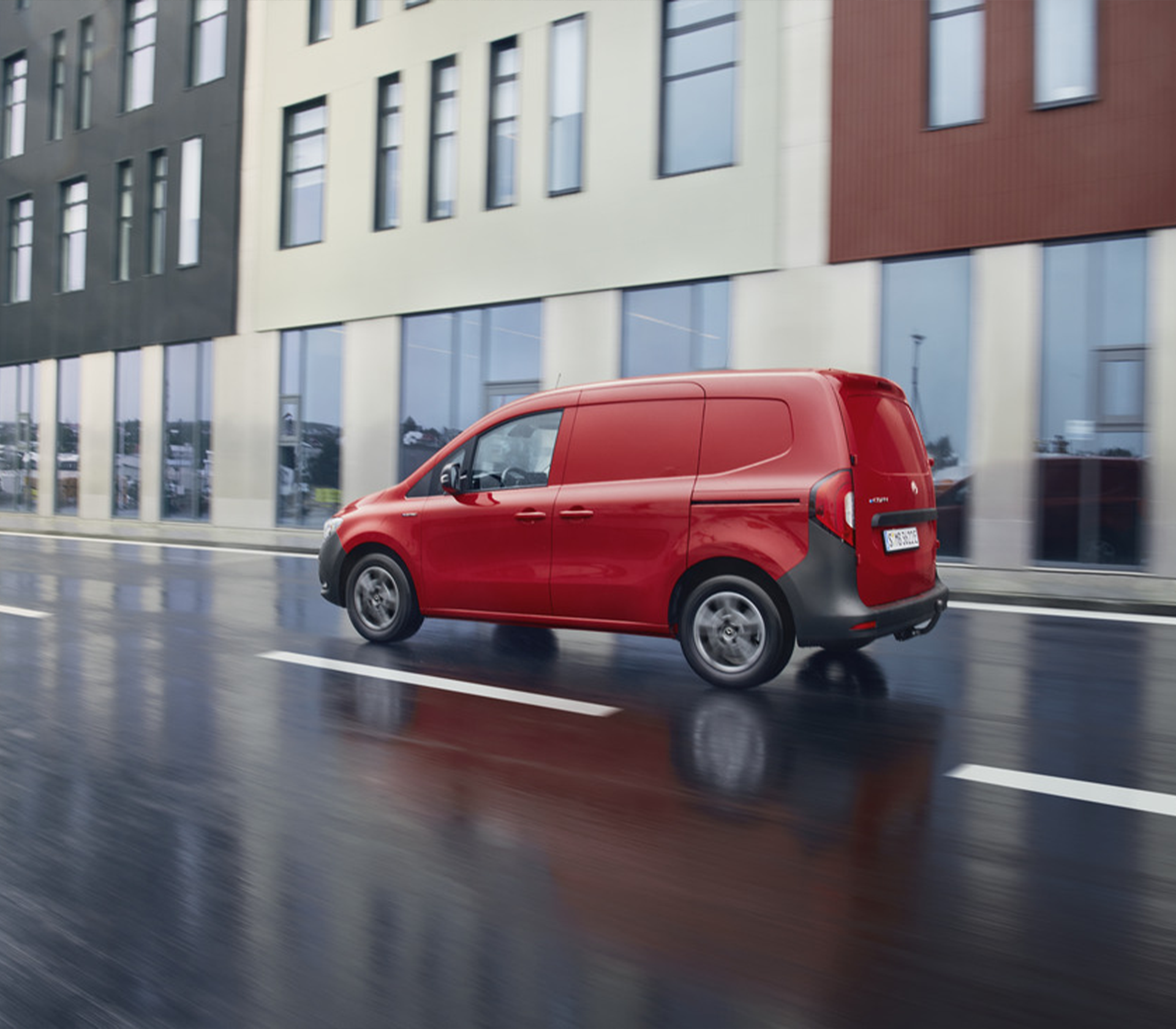 Roter Mercedes-Benz Citan fährt bei nasser Fahrbahn an modernen Bürogebäuden vorbei