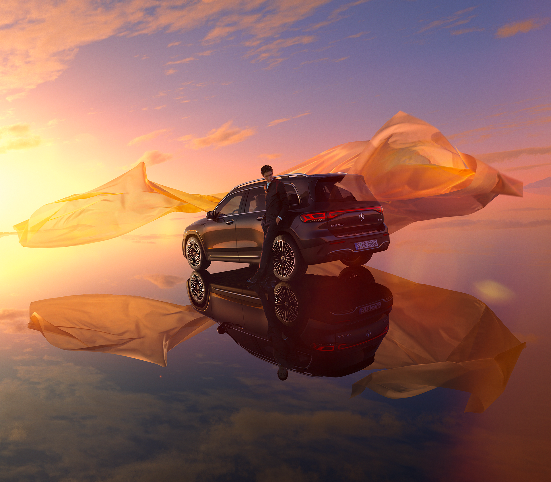 Mercedes-Benz EQB Seitenansciht mit einem großem Tuch, welches im Wind weht beim Sonnenuntergang, ein Mann lehnt gegen das Auto