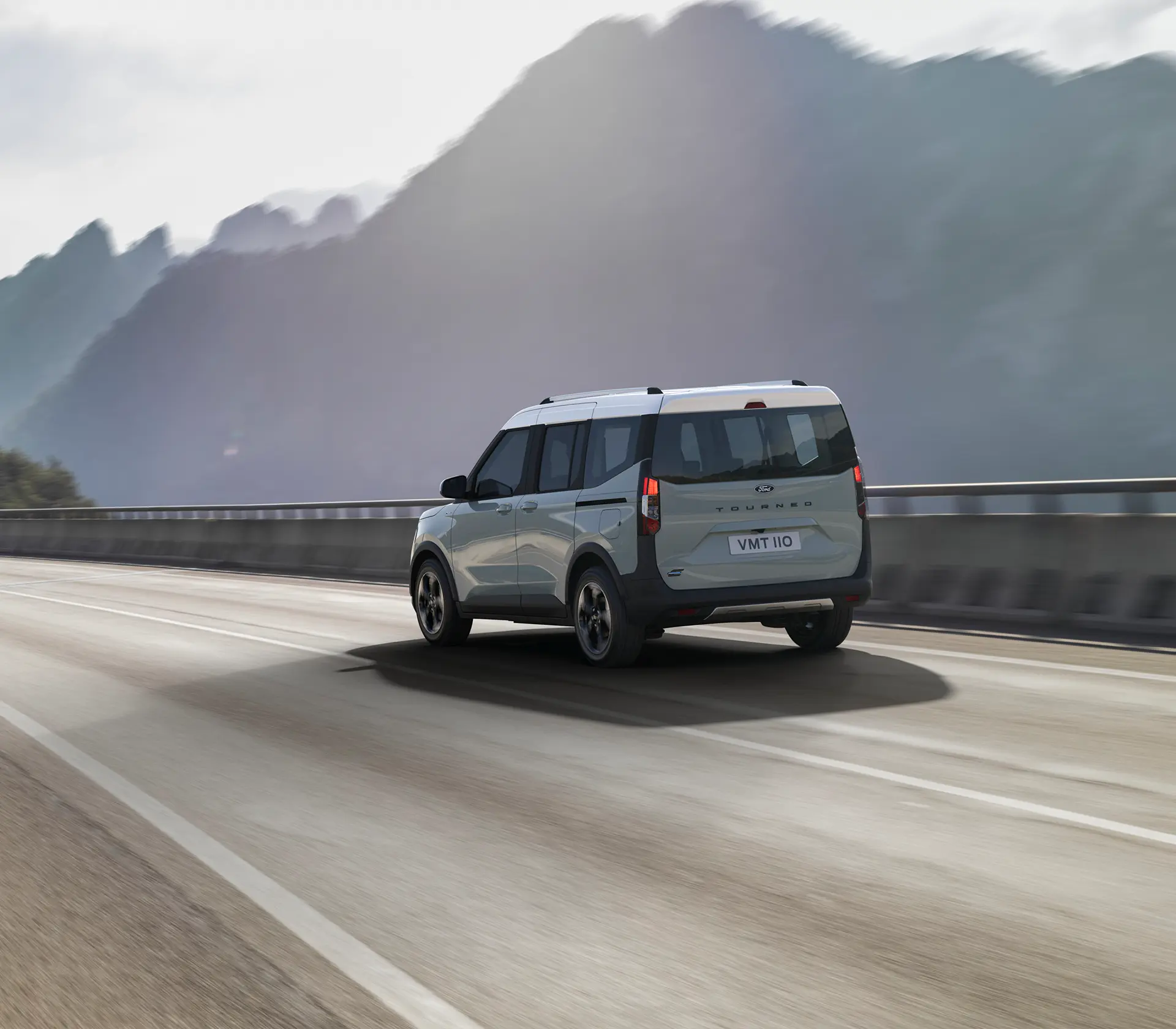 Grauer E-Tourneo Courier fährt auf der Straße mit Gebirge im Hintergrund