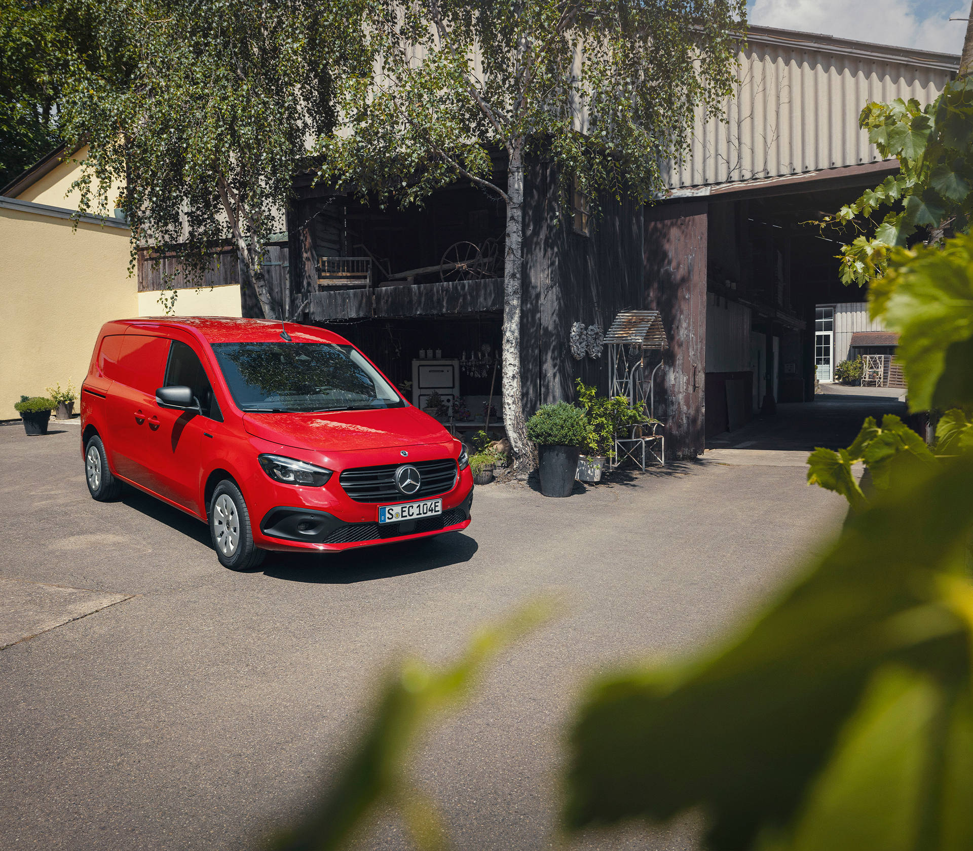 Roter Mercedes-Benz Citan steht vor einem ländlichen Gebäude im Schatten von Birken