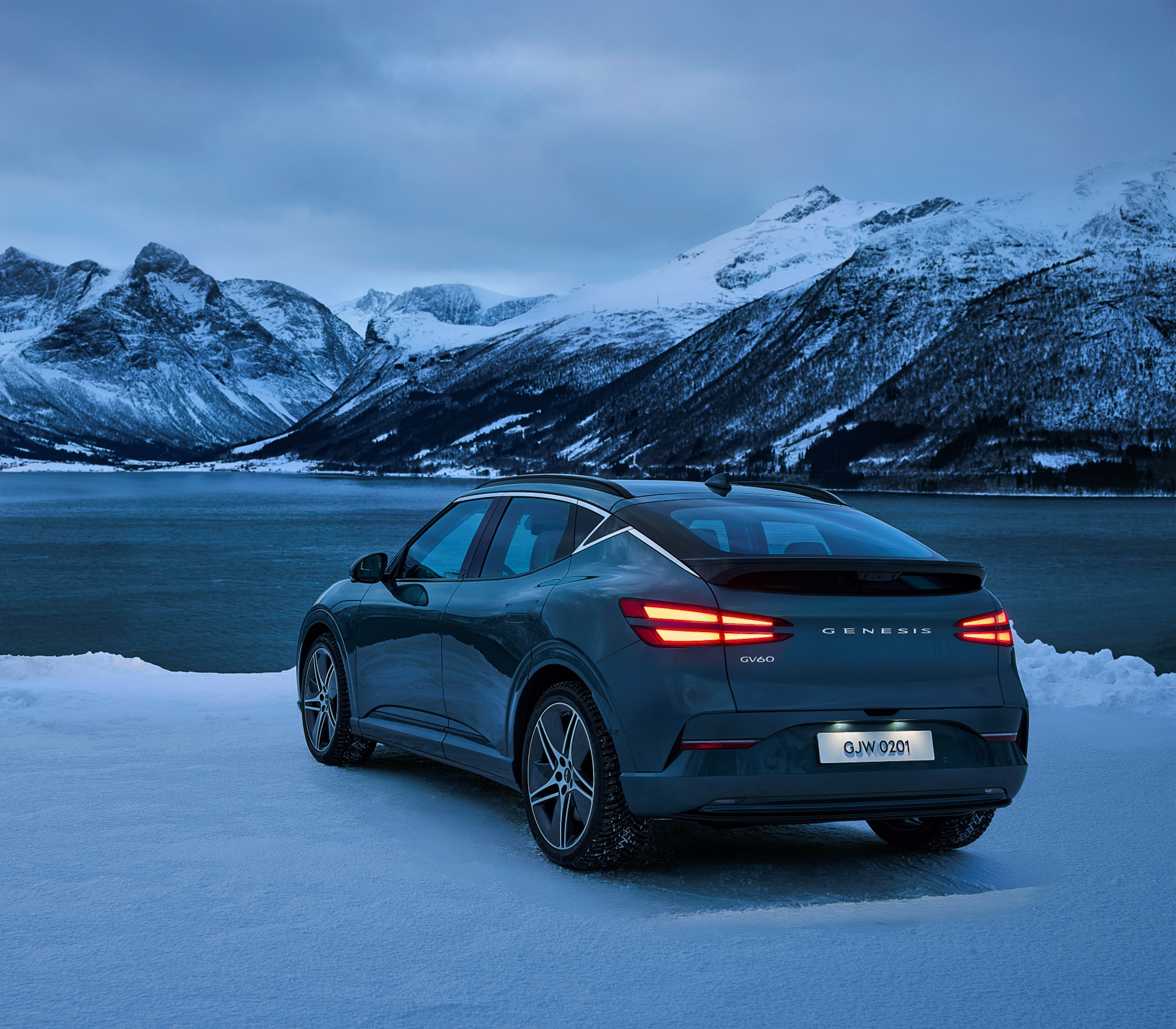 Genesis GV 60 heck- und Seitenansicht steht auf Schnee vor einem See und im Hintergrund sind schneebedeckte Berge