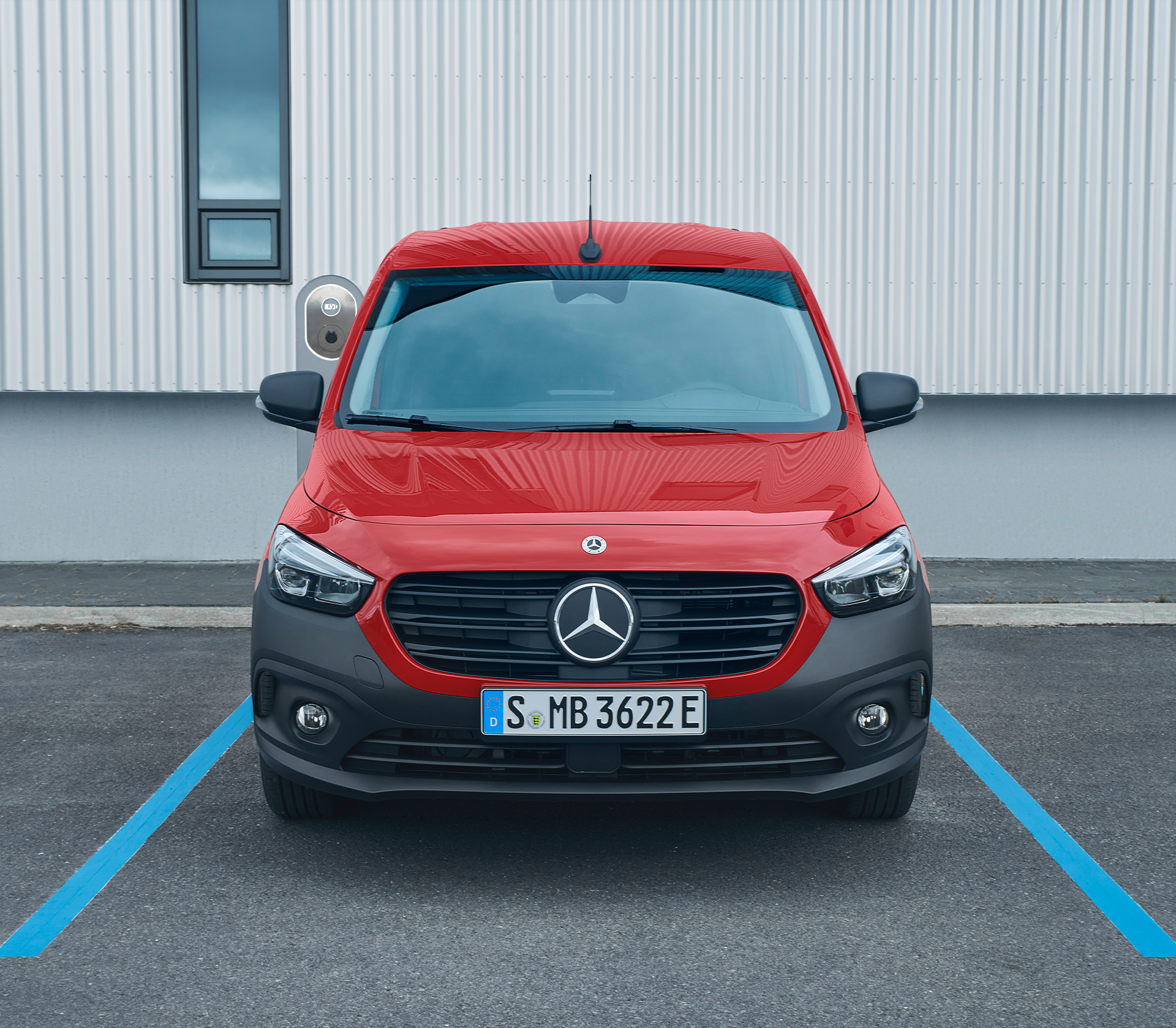 Frontalansicht eines roten Mercedes-Benz eCitan an einer Ladesäule, geparkt auf einem markierten E-Parkplatz vor moderner Industriearchitektur.