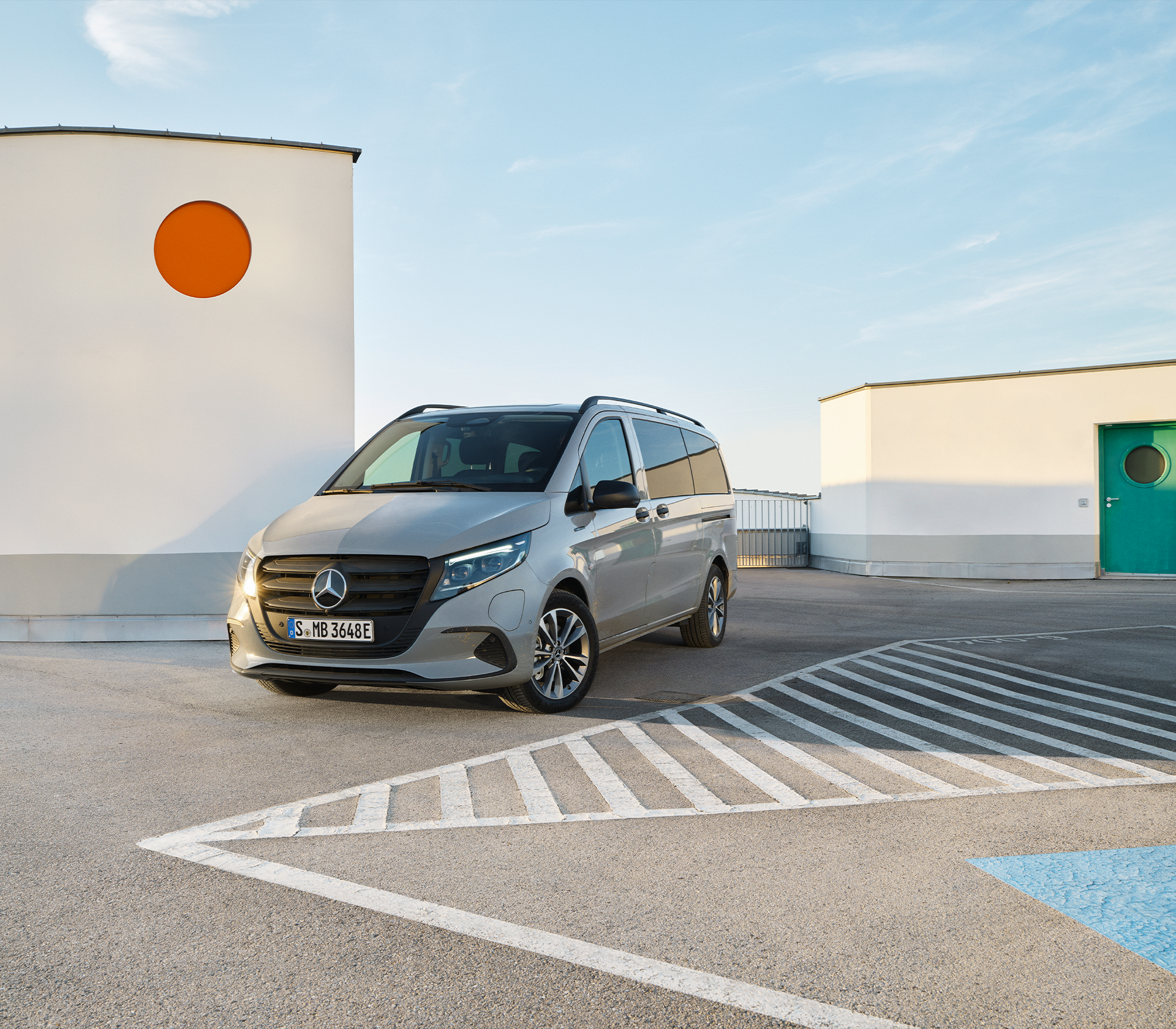 Silberner Mercedes-Benz e-Vito geparkt auf einem sonnigen Dachparkdeck vor moderner Architektur mit runder, oranger Fensteröffnung.