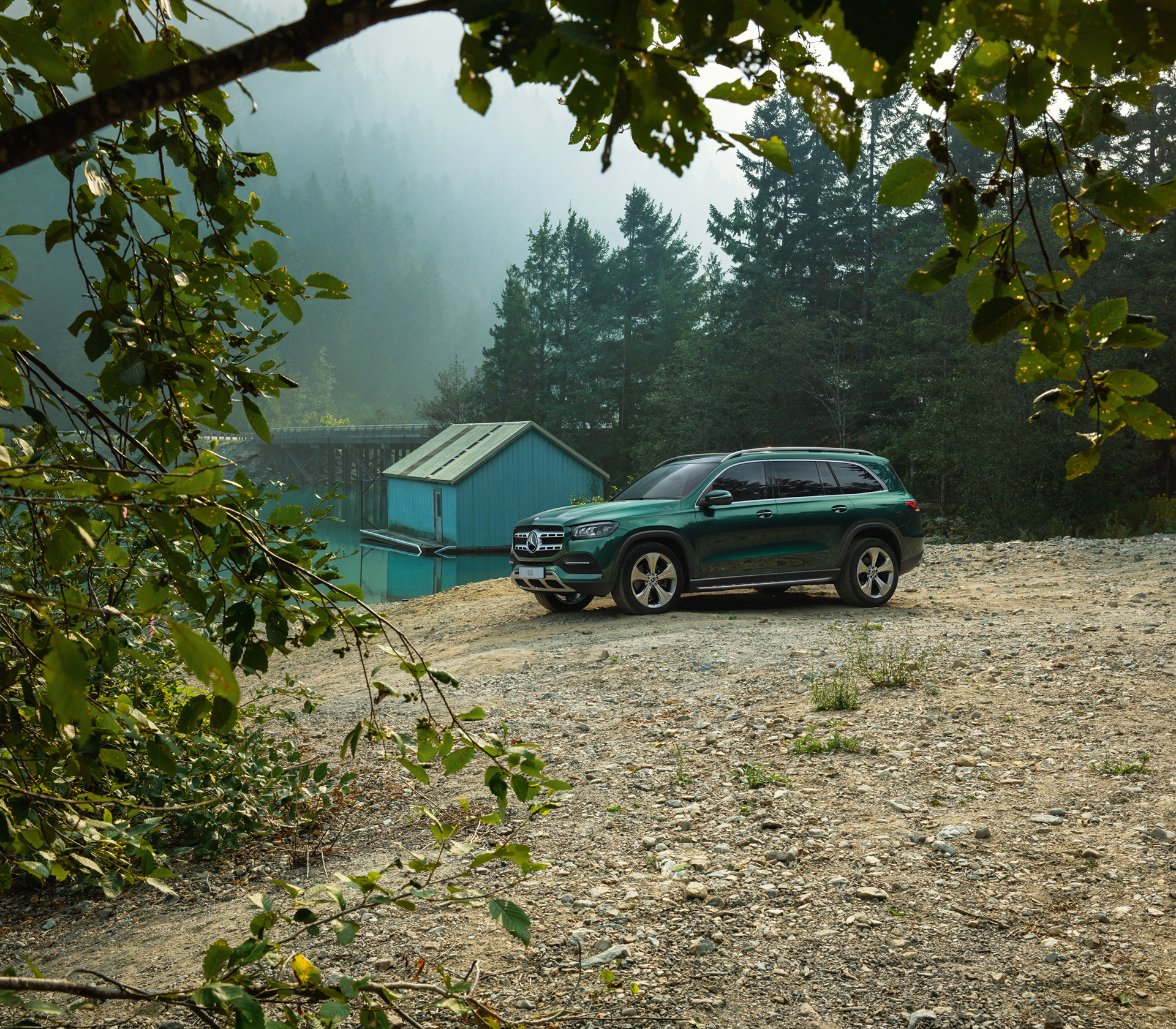 Mercedes-Benz GLS perspektive zwischen den Ästen eines Baumes auf das Auto, das neben einem See mit viel Wald und einer Hütte im Hintergrund fotografiert ist