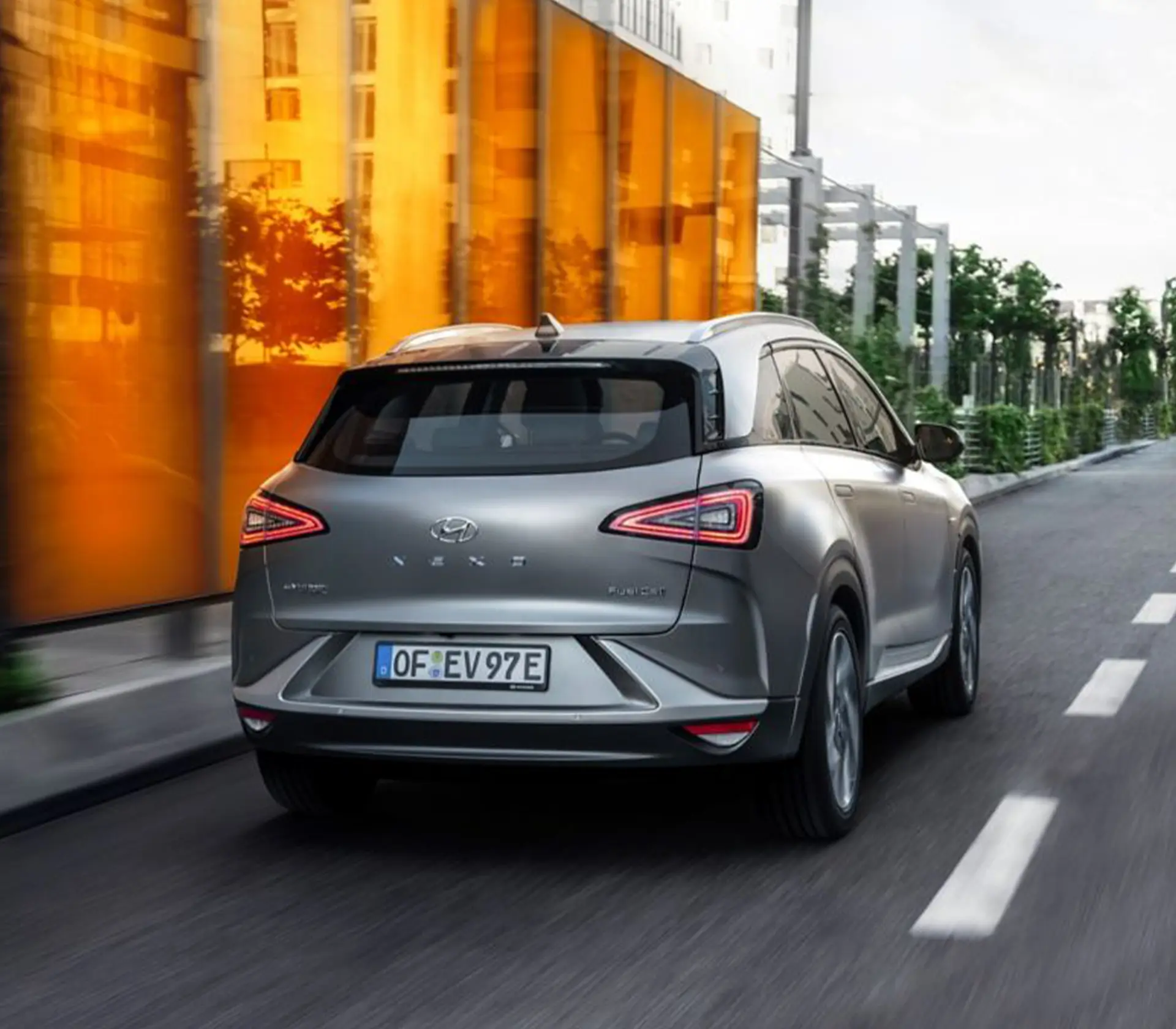 Hyundai NEXO fährt auf der Straße. Man sieht das Heck des Autos und daneben eine orangene halb durchsichtige Wand mit Gebäuden und Bäumen im Hintergrund.